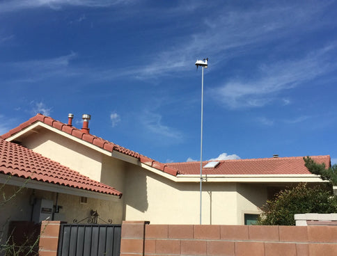 Compact white weather station installed on a tall mast above red-tiled suburban roofs under a clear blue sky